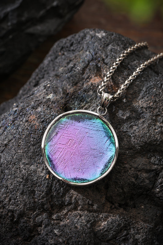 Round pendant with a purple, blue and green face in a silver-colored frame, resting on dark stone with a chain.