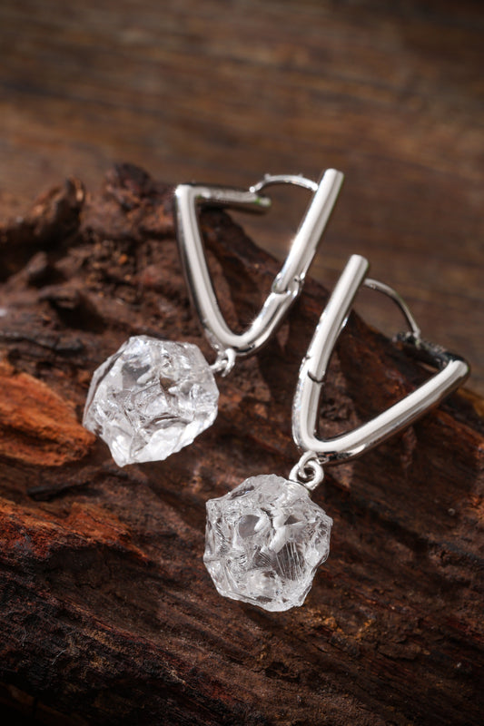 Two irregular clear quartz drops with angular silver-colored fittings resting on brown textured wood.