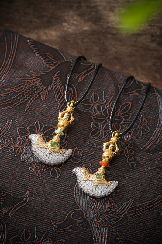 Two ornate axe-shaped pendants on black cords, with green centers and different blue and red accents, resting on dark patterned fabric.